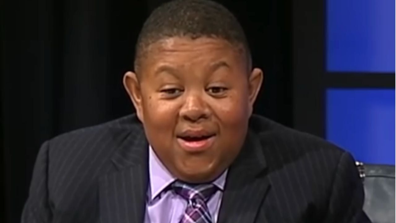 A person wearing a dark pinstripe suit, lavender shirt, and plaid tie sits indoors, smiling with a surprised or amused expression.