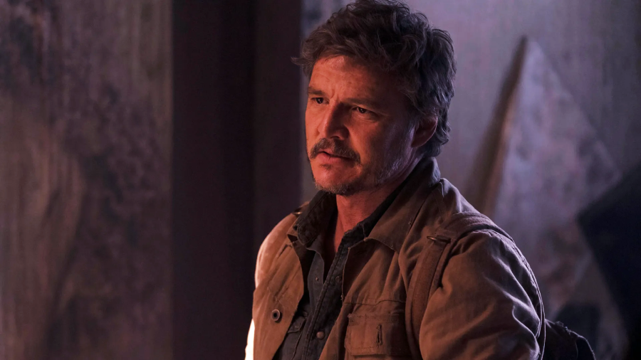 A man with short brown hair and facial hair, wearing a brown jacket over a blue shirt, stands indoors in dim, moody lighting with a serious expression on his face.