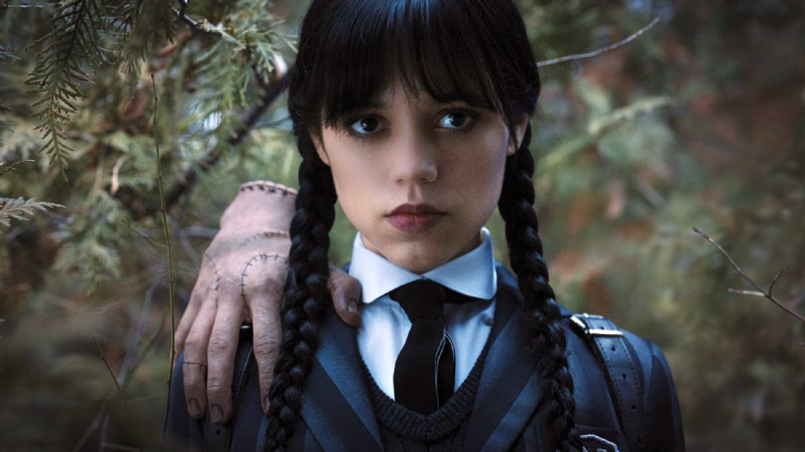 A young girl with long black braids and a serious expression stands in a forest, wearing a dark uniform and tie. A disembodied hand rests on her shoulder, partially visible among the branches.