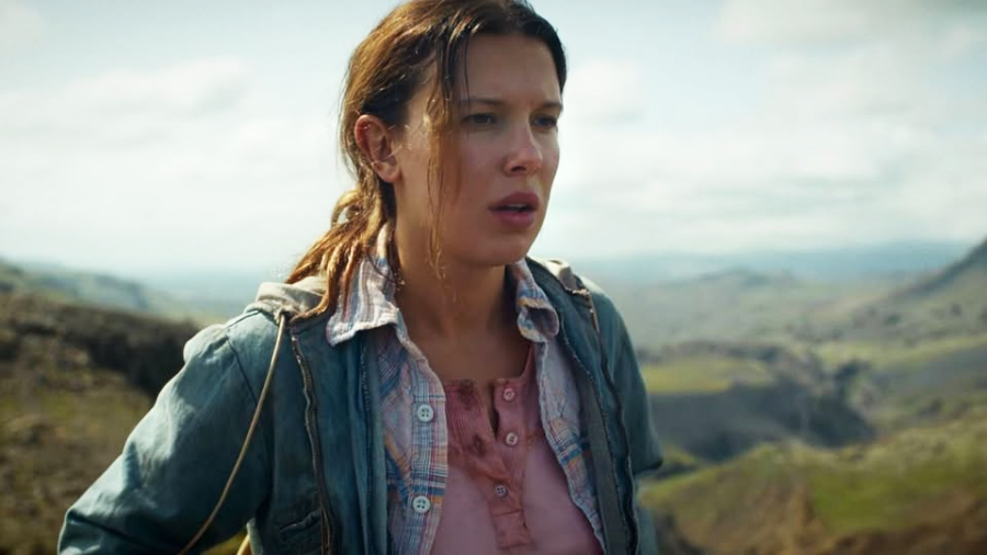 A young woman with brown hair, wearing a denim jacket over a plaid shirt, stands outdoors with a concerned expression. Rocky hills and a cloudy sky are visible in the background.
