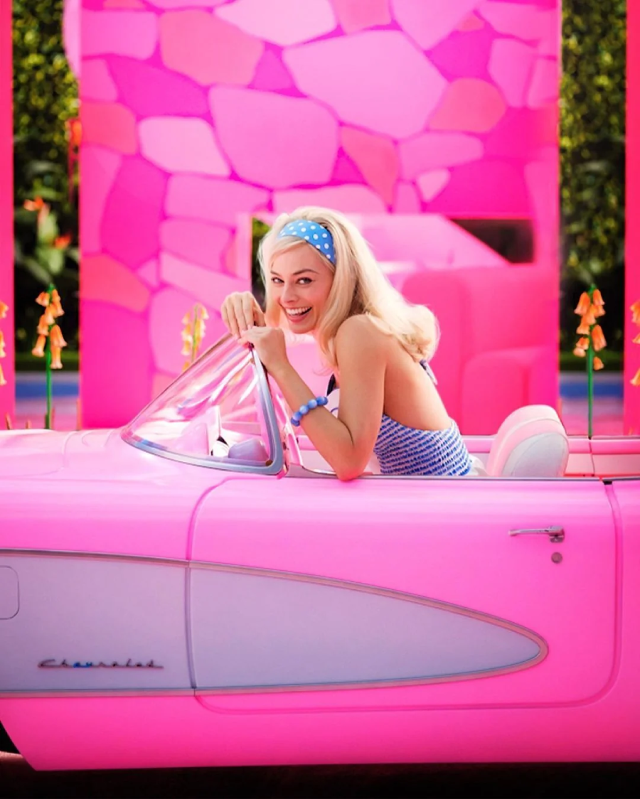 A smiling woman with blonde hair and a blue polka dot headband sits in a bright pink convertible car, wearing a blue striped top. The vibrant background features pink stone-patterned walls and colorful flowers.
