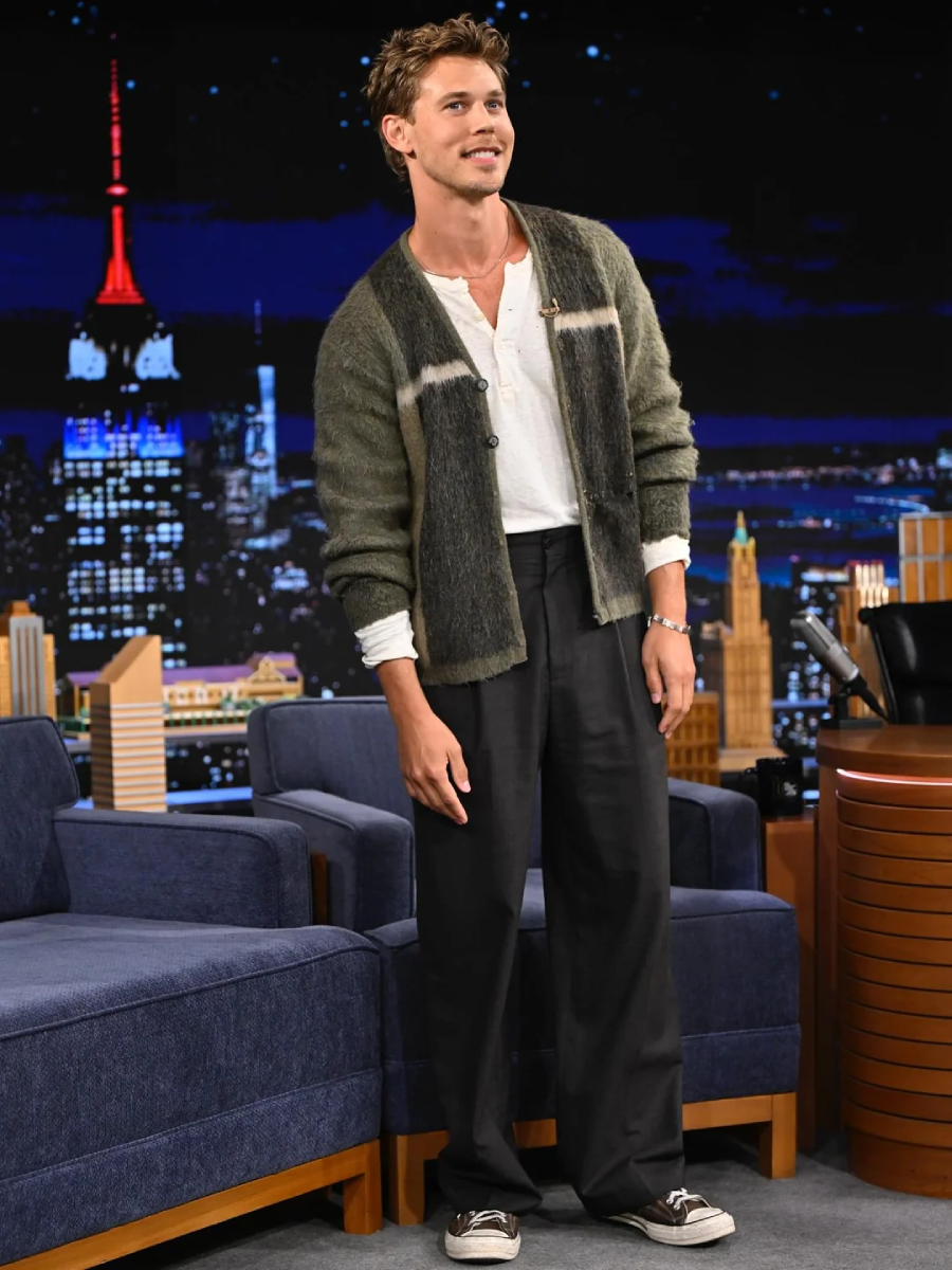 A man stands on a talk show set in front of a nighttime cityscape backdrop. He wears a white shirt, loose dark pants, a fuzzy cardigan, and sneakers, smiling casually with hands by his sides.