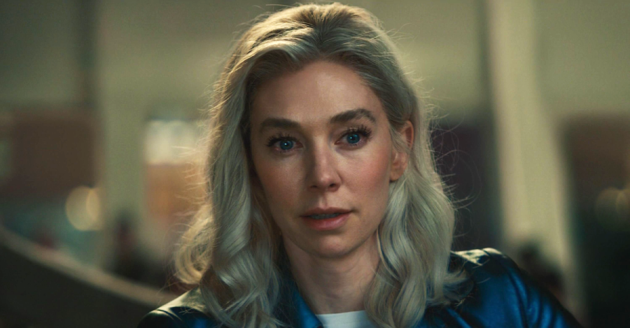 A woman with long, wavy silver-blonde hair and light skin looks forward with a serious expression. She is wearing a blue jacket and is in an indoor setting with a blurred background.