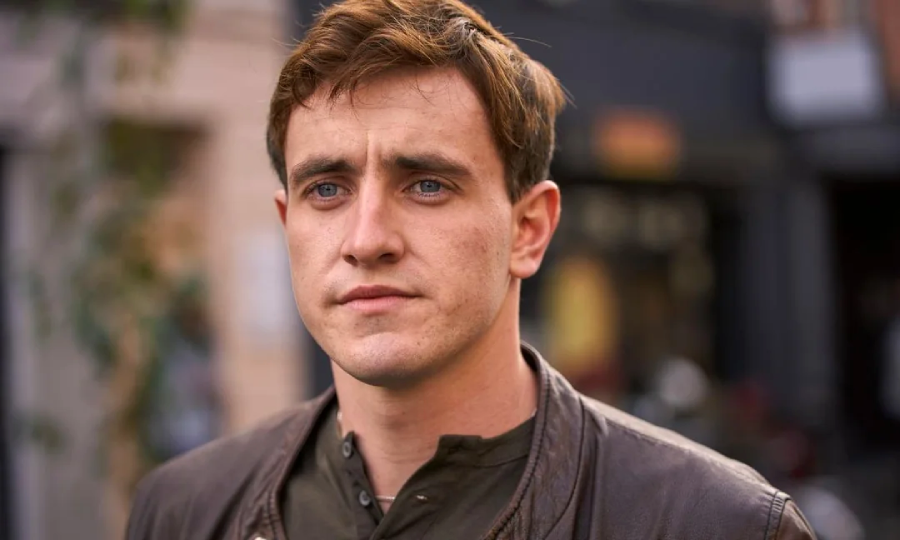 A young man with short brown hair and blue eyes stands outdoors, looking slightly to the side. He wears a brown leather jacket over a dark shirt, with a blurred urban background behind him.