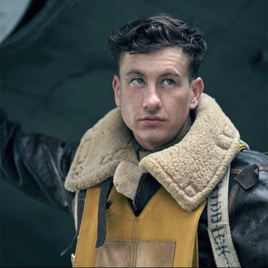 A young man wearing a World War II-era leather flight jacket with a shearling collar and yellow vest looks up thoughtfully, standing beside an aircraft.