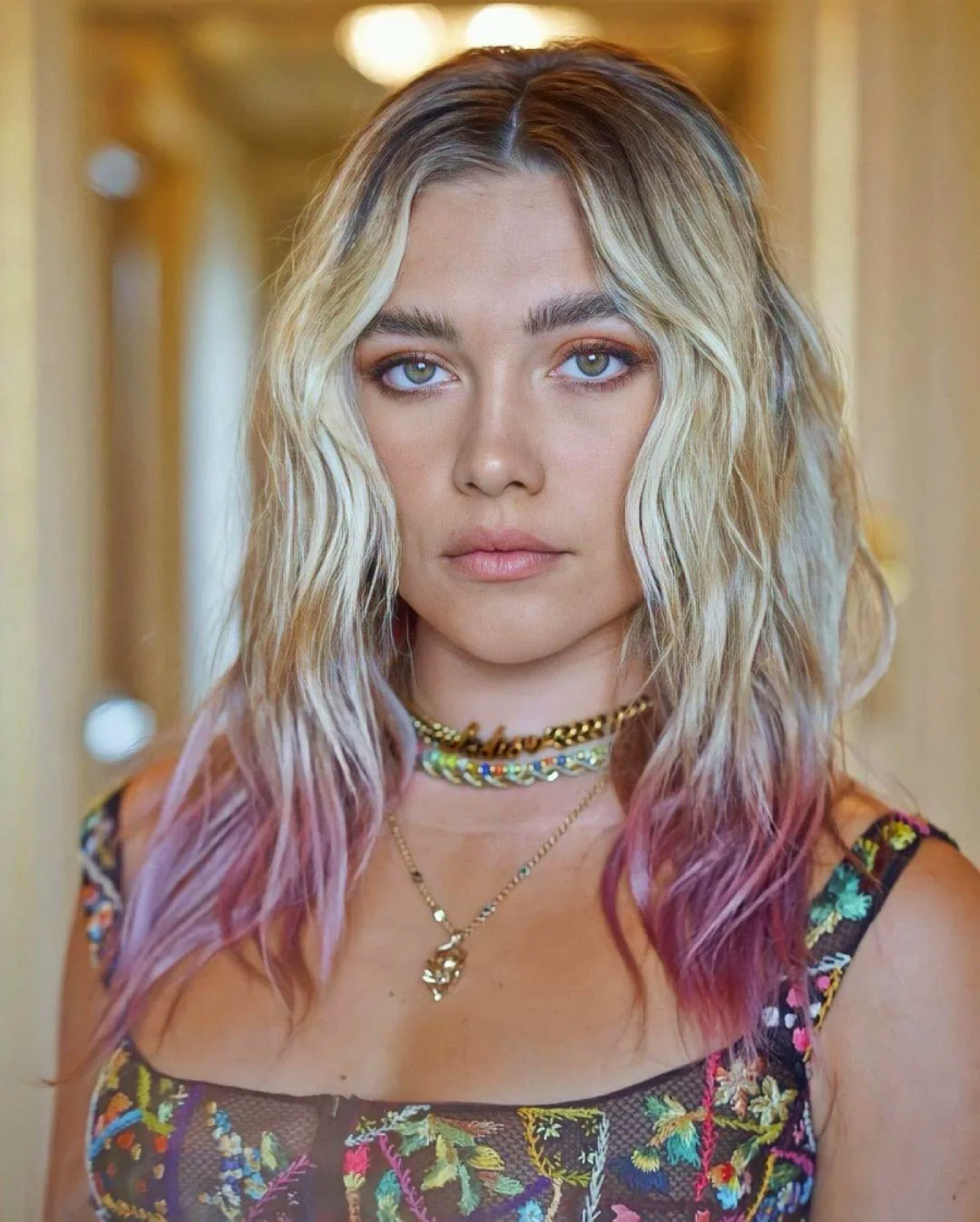 A woman with wavy blonde hair featuring purple tips, wearing layered necklaces and a floral-patterned top, stands indoors with a neutral expression and soft lighting behind her.