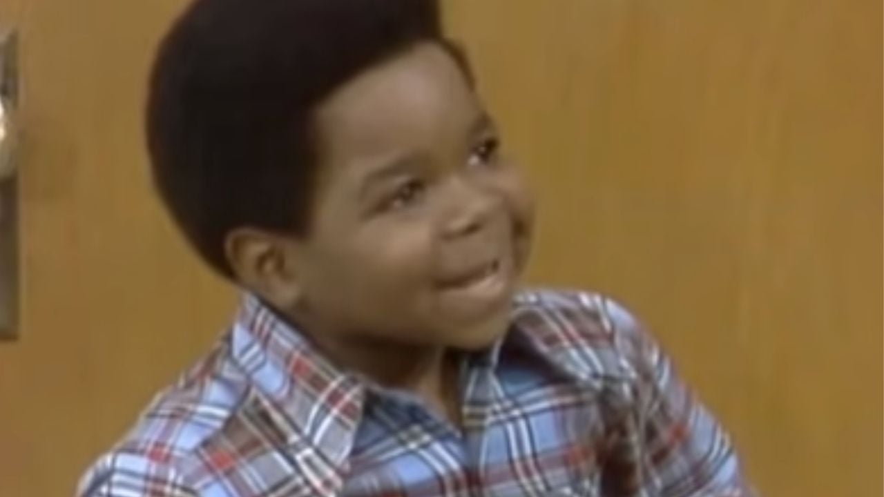 A young boy with short hair smiles and looks up. He is wearing a blue, red, and white plaid shirt and standing indoors in front of a brown background.