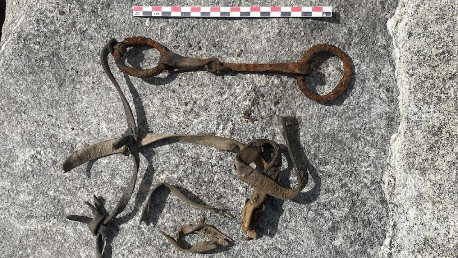 A rusty metal horse bit and worn leather straps are laid out on a gray stone surface with a measuring scale at the top for reference.
