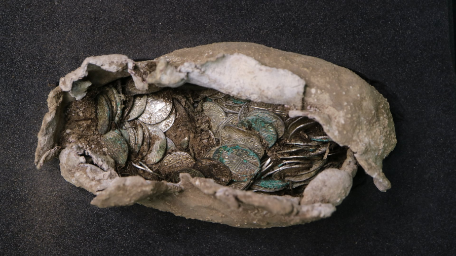 A weathered clay vessel filled with old, tarnished silver coins, some covered in green patina, sits on a dark surface.