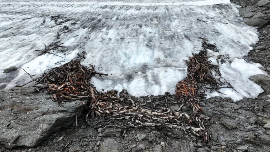 A patch of melting ice and snow on rocky ground, with clusters of exposed rusty metal sticks and debris arranged at the ice’s edge.