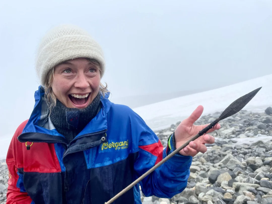 A person in a blue and red jacket and white beanie smiles excitedly while holding an ancient spearhead on a rocky, snow-covered mountain landscape.