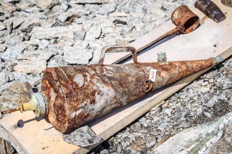 A large, rusted, cone-shaped metal object with a handle and a numbered sticker rests on a wooden plank among scattered rocks. A smaller, similarly rusted cylindrical object lies nearby.