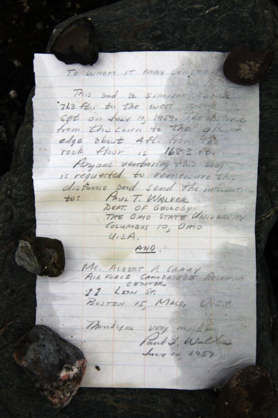 A handwritten letter on lined paper, weighed down by rocks at the corners, rests on a dark surface. The letter includes names, addresses, dates, and a brief message about a bottle sent from Ohio.
