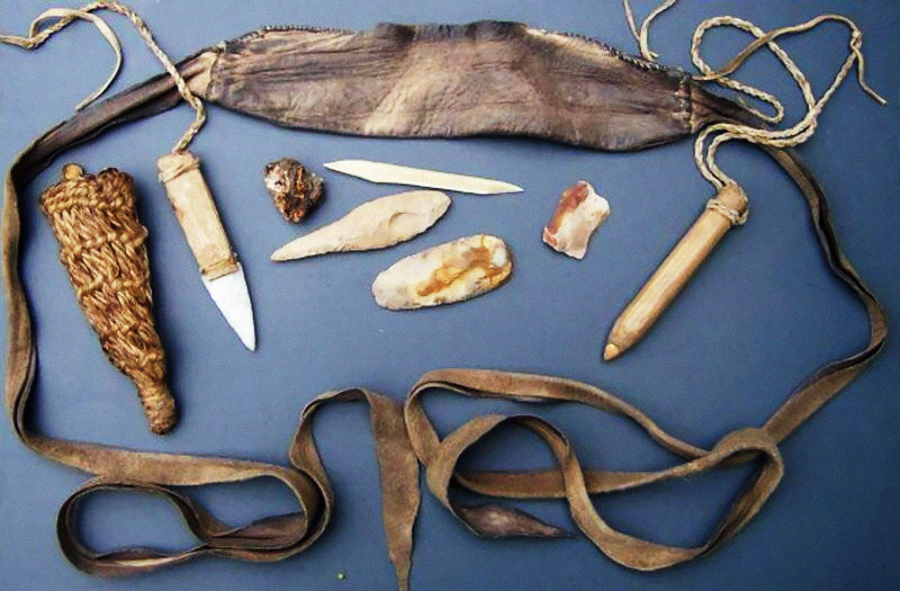 A set of ancient or prehistoric tools including stone blades, bone implements, a woven sheath, and a leather pouch with straps, arranged on a flat surface.
