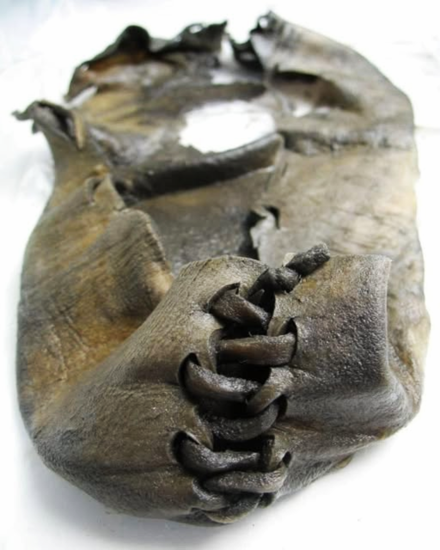 A close-up of an ancient leather shoe, showing a rough, dark surface with visible stitching made from leather thongs along the front. The shoe appears worn and weathered, likely an archaeological artifact.
