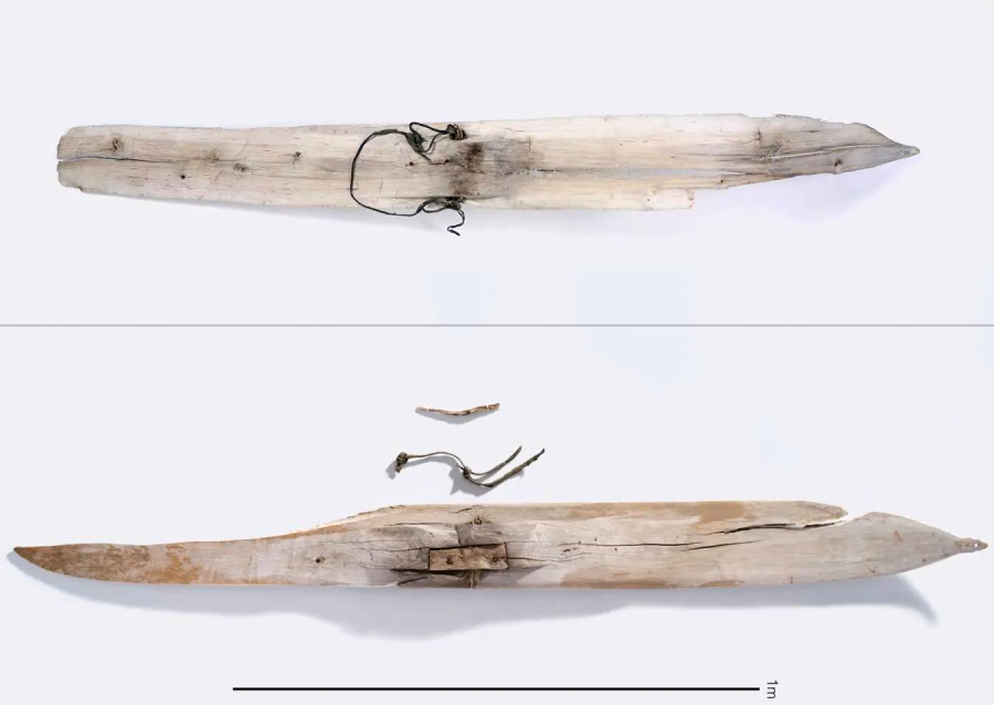 Two views of an old, weathered wooden ski with a pointed tip and a basic leather binding attached; the bottom view also shows a loose piece of wood and string, with a scale indicating length.