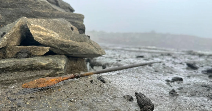 A rusty iron spearhead with a long shaft lies on wet, rocky ground near a pile of flat stones, with a foggy, blurred landscape in the background.