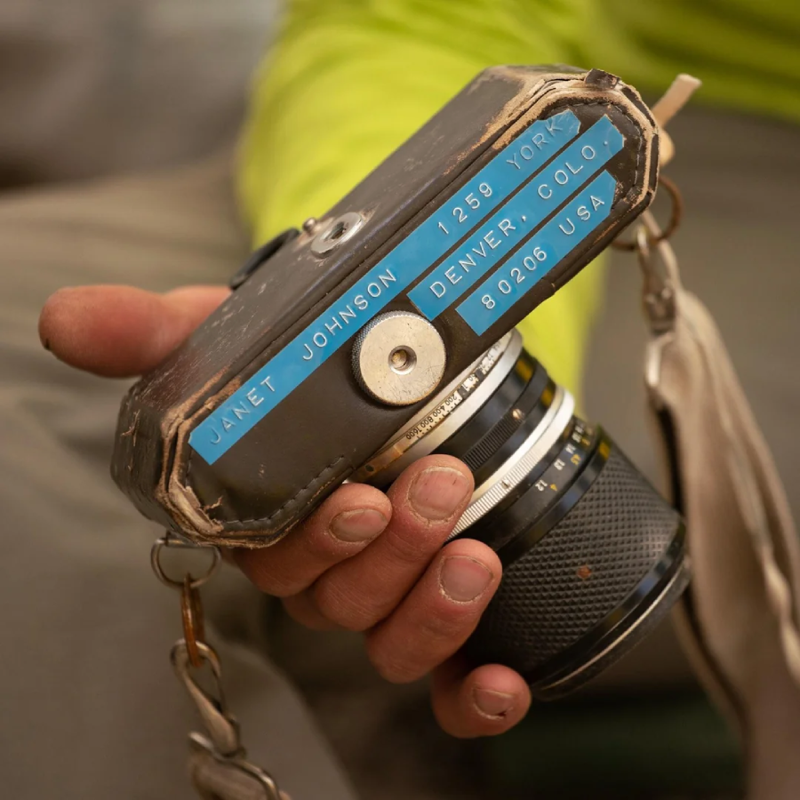 A person holds a worn camera with blue labels reading "Janet Johnson, 1258 York, Denver, Colo, 80206 USA." The camera shows signs of heavy use and has a beige strap attached.