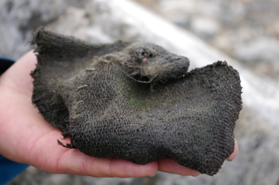 A close-up of a hand holding a worn, dark-colored, rectangular piece of coarse fabric, possibly old or weathered clothing, against a blurred outdoor background.
