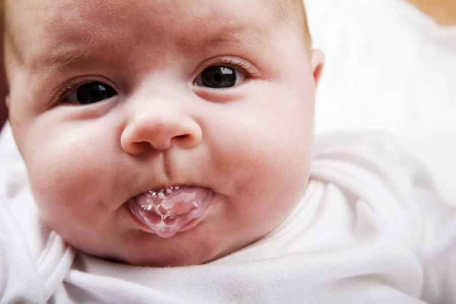 Close-up of a baby with big eyes and chubby cheeks, sticking out their tongue with bubbles of saliva on it, wearing a white outfit.