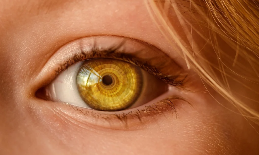 Close-up of a human eye with a golden, mechanical-looking iris featuring concentric circles, giving a futuristic or robotic appearance. Strands of blonde hair partially frame the upper right of the image.