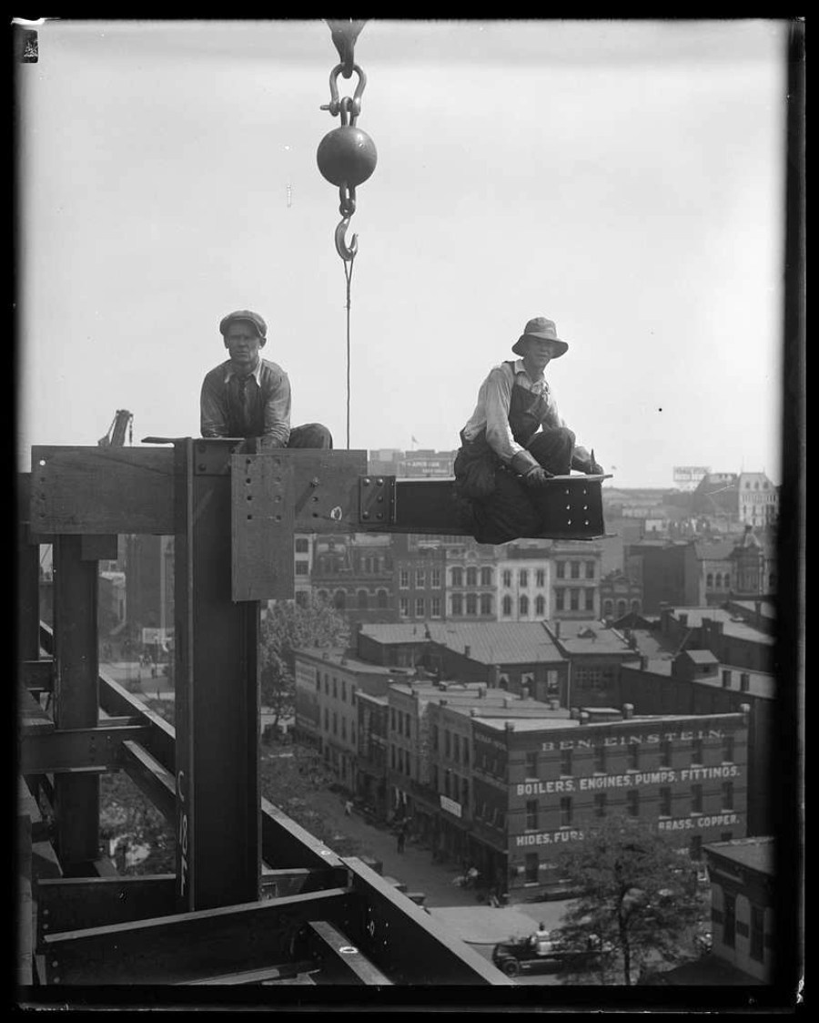 Two construction workers sit on a steel beam high above a city street, with a crane hook hanging nearby. City buildings and signs for industrial businesses are visible in the background.