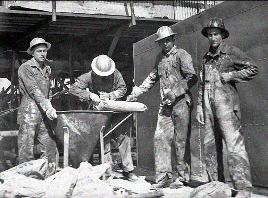 Four construction workers in hard hats and work clothes stand around a large metal container at a building site. One worker handles a bag, while others pose or watch, surrounded by construction materials.