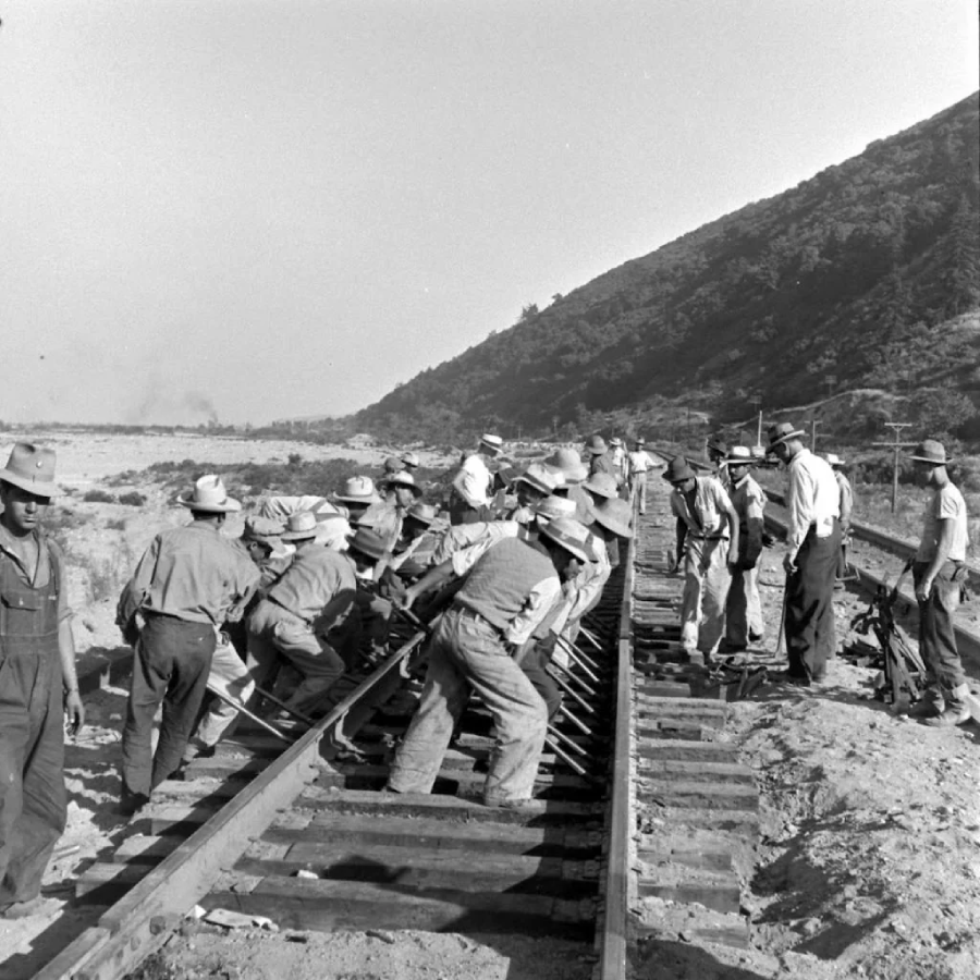 A group of workers wearing hats and work clothes use tools to lay or adjust railroad tracks in a rural, hilly area. The scene is outdoors, with a dirt embankment and a mountain in the background.