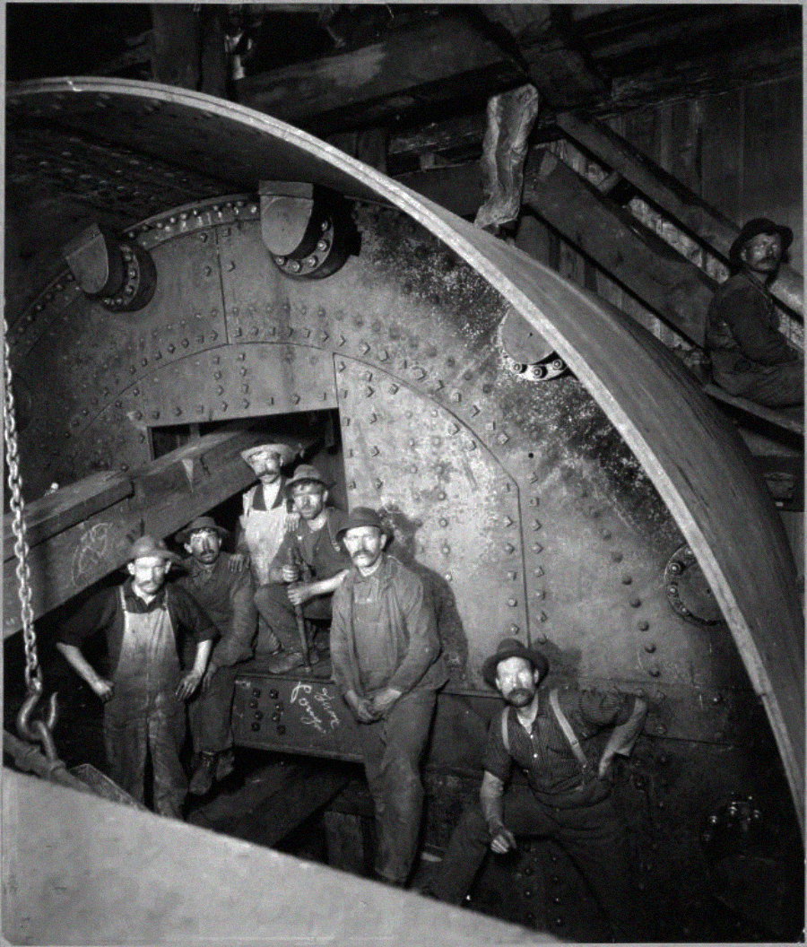 Black-and-white photo of seven men in work clothes and hats standing inside and around a giant industrial machine, likely in a factory or mill setting, with large metal bolts and beams visible.