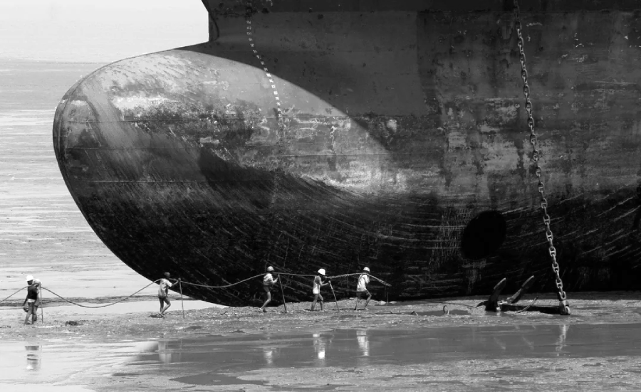Five workers in helmets pull a thick rope across a muddy shore in front of the massive hull of a beached ship. The ship's dark, weathered metal dominates the background. The scene is in black and white.