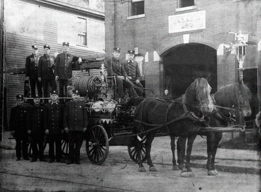 Vintage black-and-white photo of firefighters in uniform posing with a horse-drawn fire engine in front of a brick firehouse labeled "Engine 1." Two horses are harnessed to the fire engine.