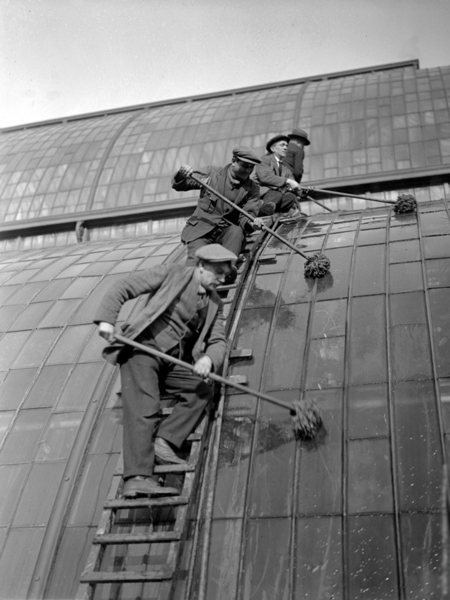 Three men in work clothes and caps clean the curved glass roof of a building using long-handled brushes while standing on ladders and scaffolding. The background shows more glass panels.
