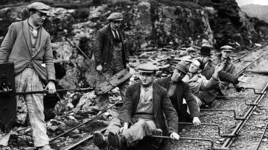 A group of men in work clothes and hats sit on a railway track using hand levers, with rocky terrain in the background, suggesting manual labor or mining work in an early 20th-century setting.