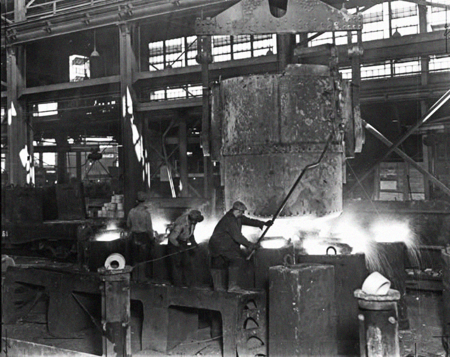 Three workers in an industrial foundry pour molten metal from a large ladle into molds, surrounded by machinery and beams inside a large factory building. Bright sparks and glowing metal are visible.