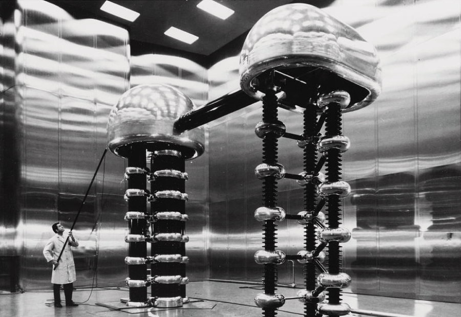 A person in a lab coat stands next to two large, metallic, dome-shaped electrical machines connected by a thick cable inside a shiny, industrial room with reflective walls.