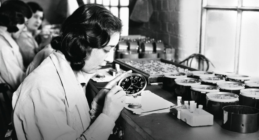 A woman in a lab coat paints numbers onto a clock face at a workbench, surrounded by various clock faces and tools. Other women work in the background near a window.