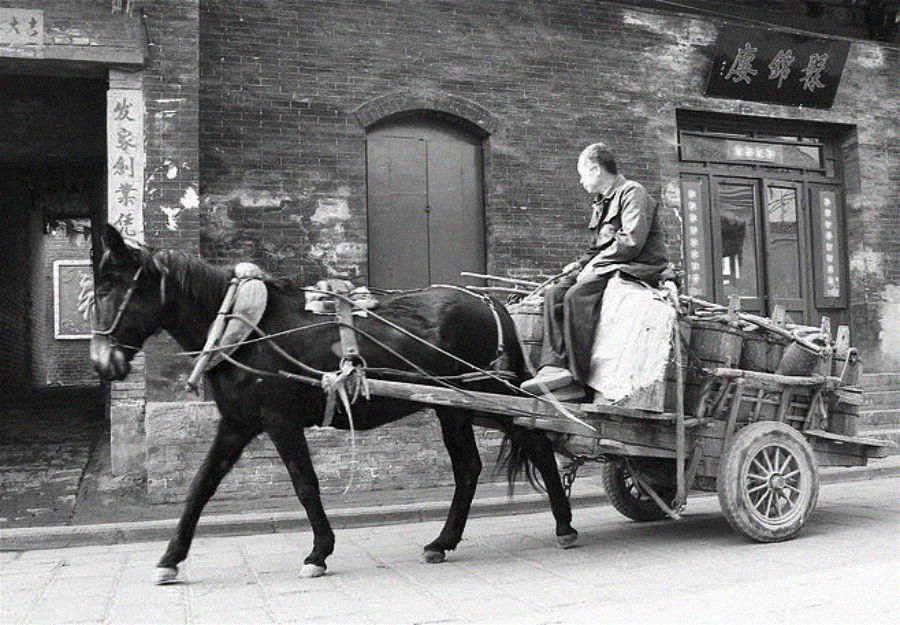 A person sits on a wooden cart pulled by a horse along a street lined with old brick buildings, with Chinese characters visible on storefront signs and posters.