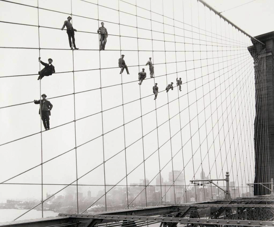 Several men in work clothes climb and pose on the large vertical cables of a suspension bridge, high above the city skyline in the background. The scene appears to be from the early 20th century.