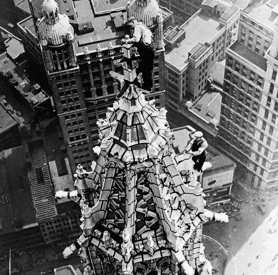 Two workers stand on the intricate spire of a tall skyscraper, high above a city with many buildings below. The perspective shows the great height and the detailed architecture of the spire.