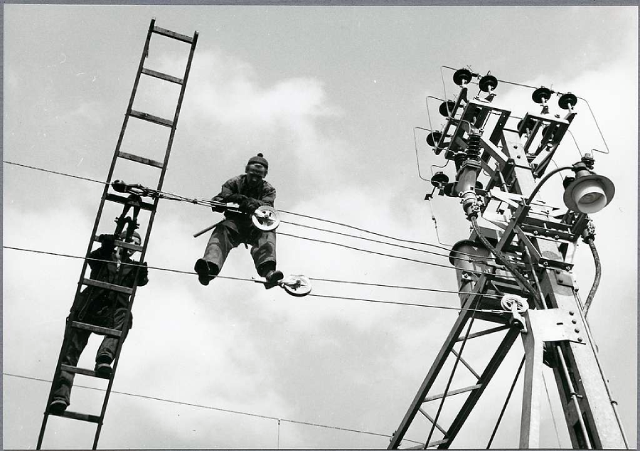 Two utility workers suspended on power lines, one sitting on a wire and the other on a ladder, are performing maintenance near a tall electricity pole against a cloudy sky.