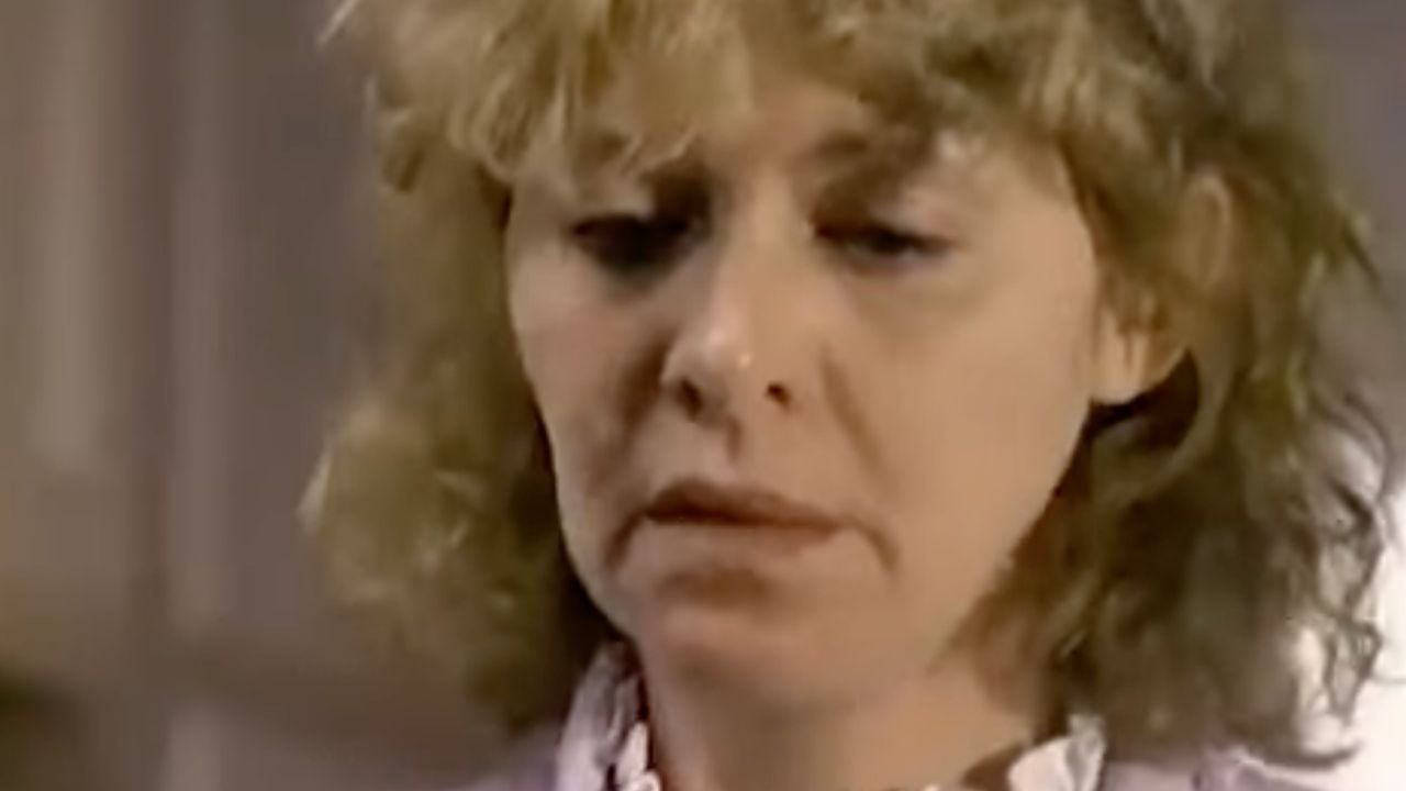 A close-up of a woman with curly blonde hair looking downward with a serious expression, wearing a light-colored top with a ruffled collar.