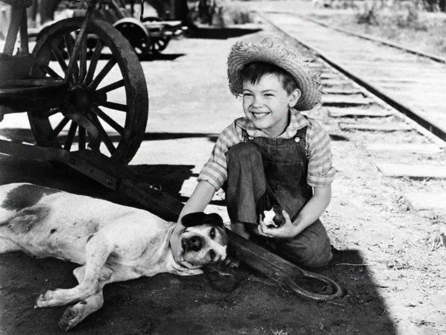 A young boy in overalls and a straw hat smiles while petting a dog lying on the ground beside him, near wagon wheels and railroad tracks, in a black-and-white vintage setting.