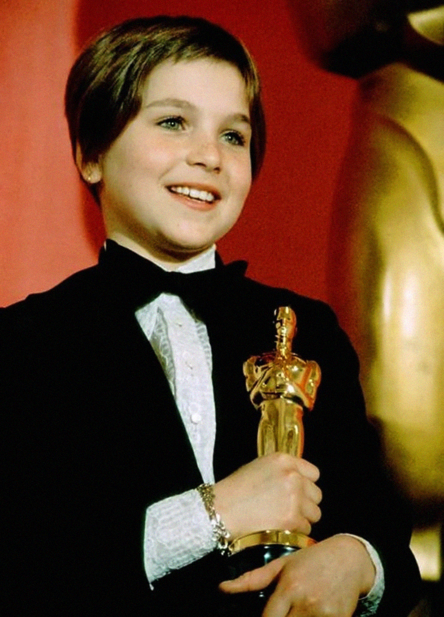 A young child with short hair, dressed in a tuxedo, smiles while holding an Oscar statuette in front of a red background and a golden statue.