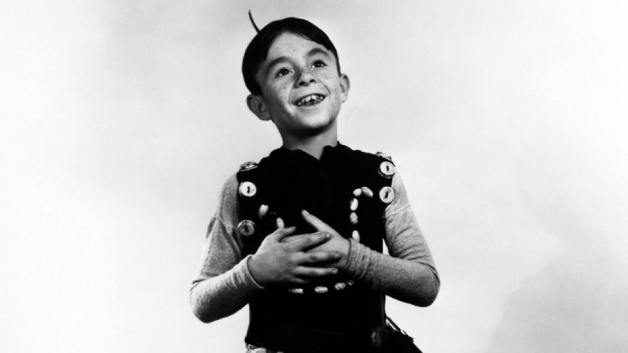 A young boy with a missing front tooth smiles and looks upward, wearing a light shirt and a vest decorated with buttons, standing against a plain white background.