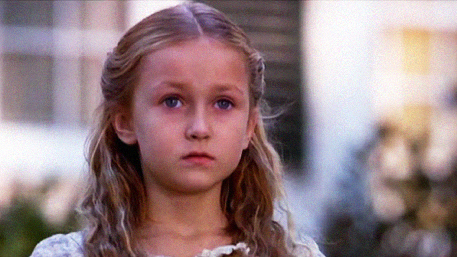A young girl with long, wavy blonde hair and light eyes looks ahead with a serious, slightly sad expression. She is outdoors, with a blurred background of greenery and a white building.