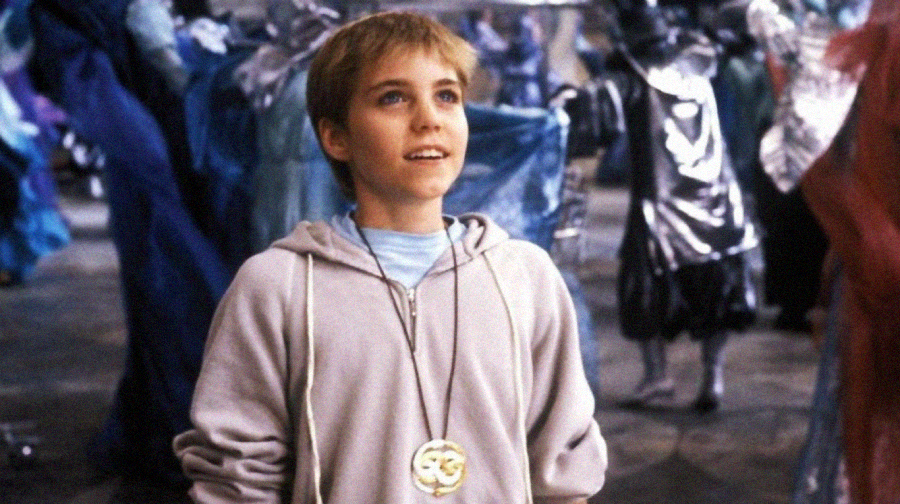 A young boy with short blonde hair wearing a grey hoodie and a large round pendant smiles while looking up, surrounded by people in Columbina masks and colorful costumes at a lively event.