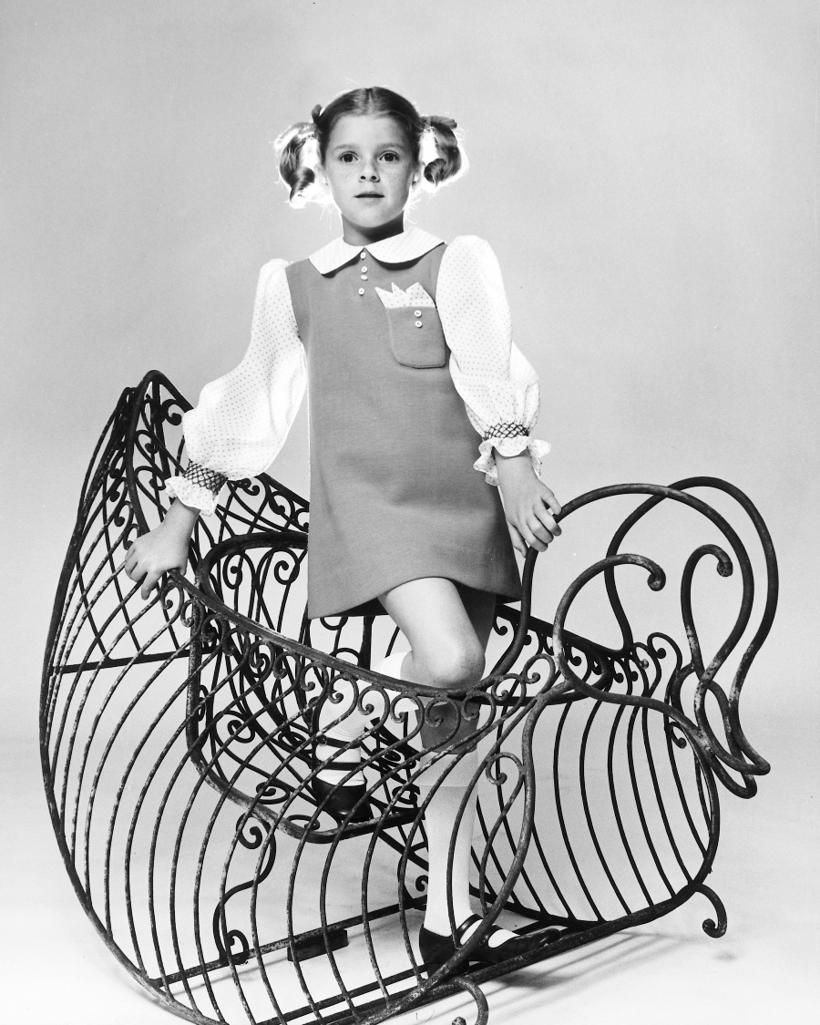 A young girl with pigtails stands confidently on a large, ornate wire rocking horse. She wears a short-sleeved dress with a white collar and cuffs, and looks directly at the camera in a studio setting.