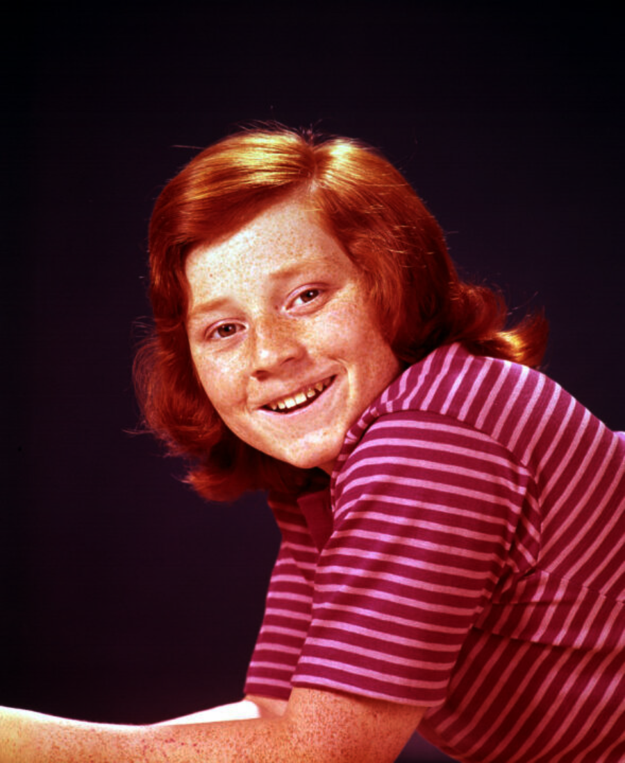 A smiling child with red hair and freckles, wearing a short-sleeve, purple and pink striped shirt, poses against a dark background.