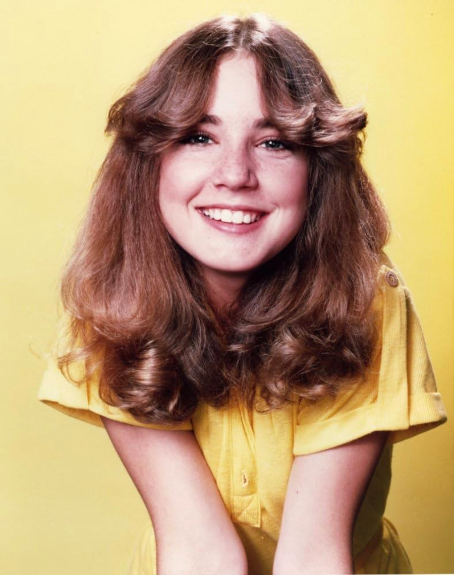 A young woman with long, wavy brown hair and bangs smiles brightly. She is wearing a yellow shirt and is posed against a solid yellow background.