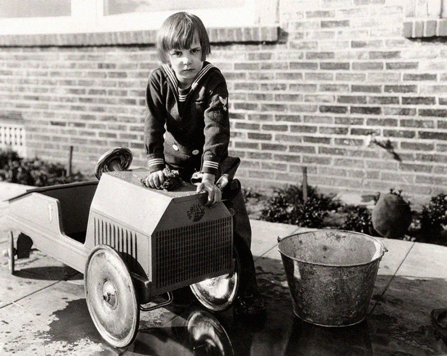 A young child in a sailor outfit washes a toy car with a metal bucket of water beside them on a paved surface, with a brick building in the background.